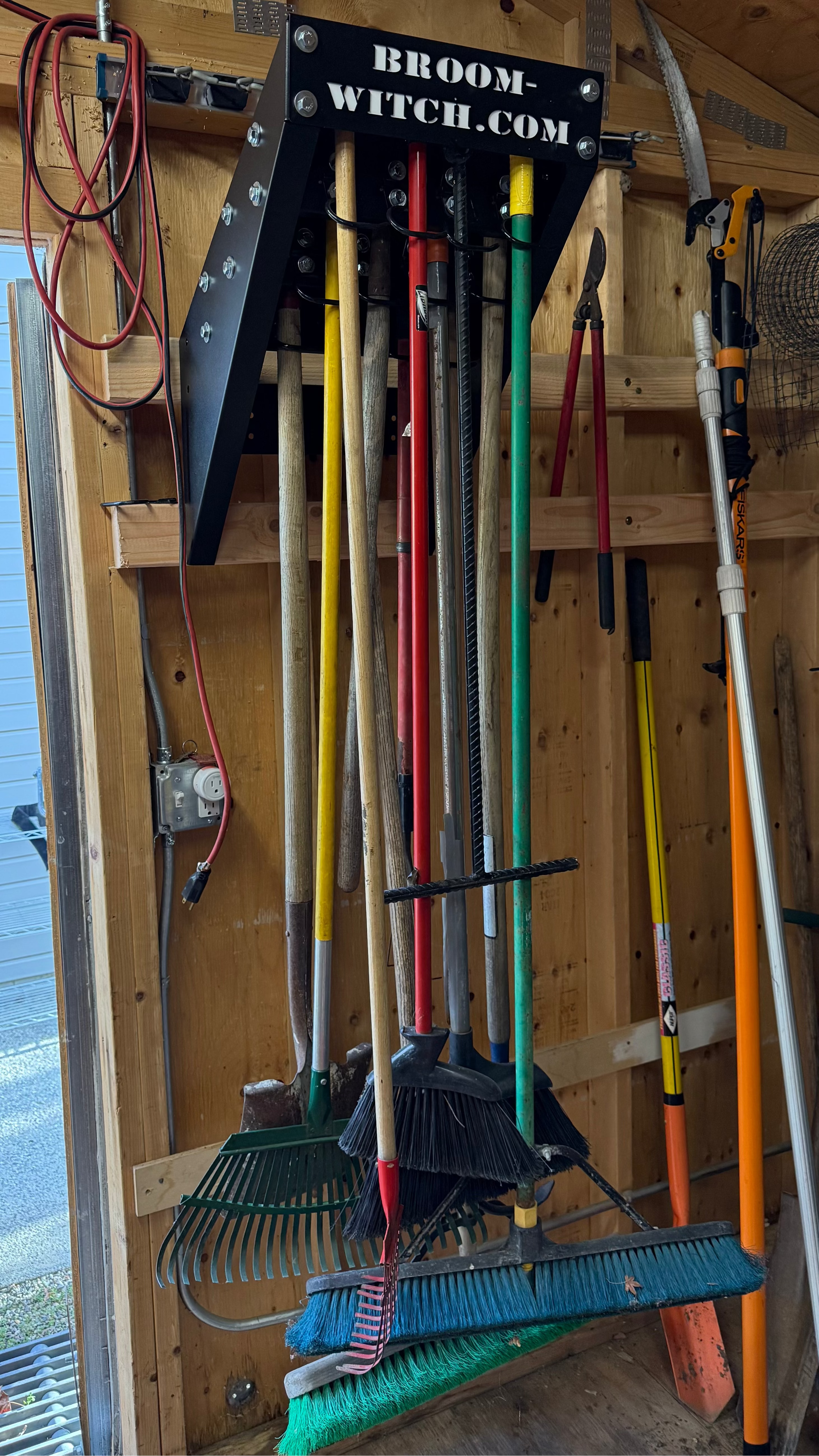 Collection of brooms and rakes hanging on a wooden wall with 'BroomWitch.com' branding.